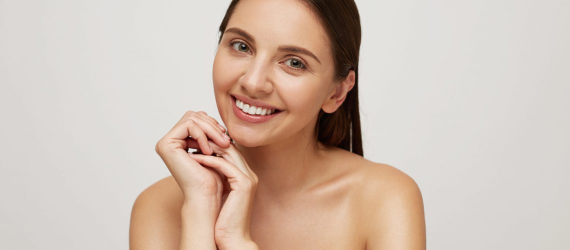 Girl with perfect healthy fresh skin looks camera as in the mirror, smiling, palms folded under the chin, very glad and satisfied with result, head and naked shoulders, over white background