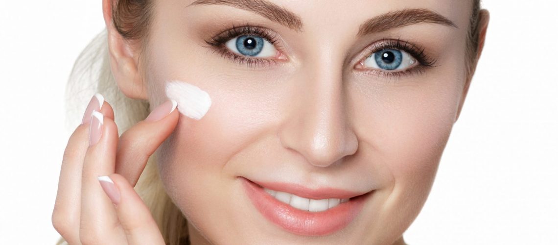 Beautiful young blonde woman applying moisturizing face cream. Woman portrait over white background. Skincare, beauty treatment, cosmetology, spa salon and skin treatment concept. Studio shot